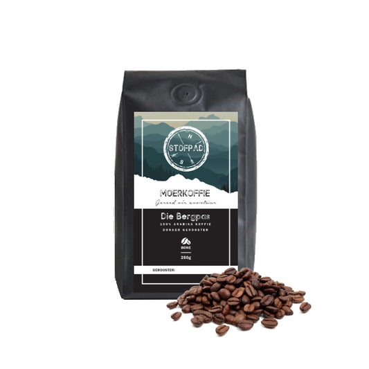 A pack of Die Bergpas Moerkoffie coffee beans with a dark roast label, indicating a strong body and caramel flavors. The beans are displayed in front of the packaging.