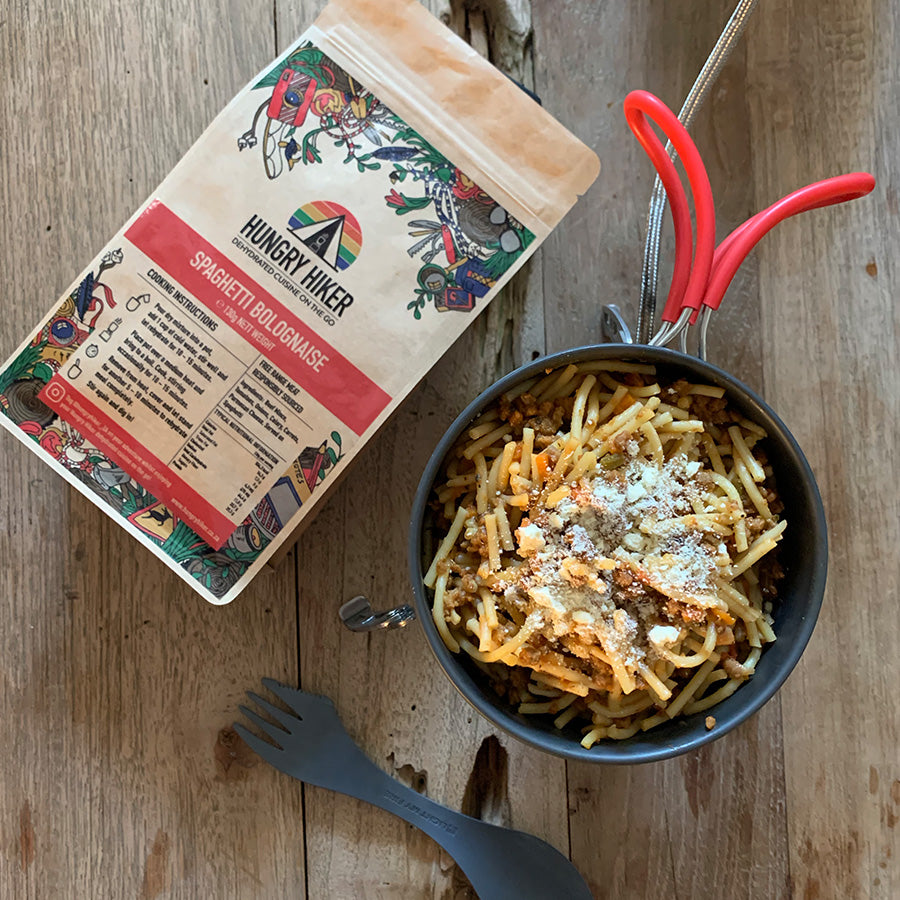 Dehydrated Spaghetti Bolognese hiking food packet with the product prepared in a pot on a wooden surface, showing the spaghetti with sauce and cheese on top.