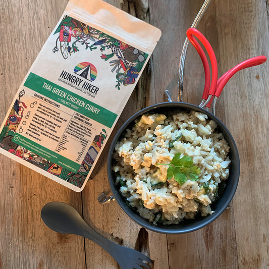 A package of Thai Green Curry dehydrated food next to a pot of cooked rice with chopped herbs on top, on a wooden surface with a red spoon and a fork.