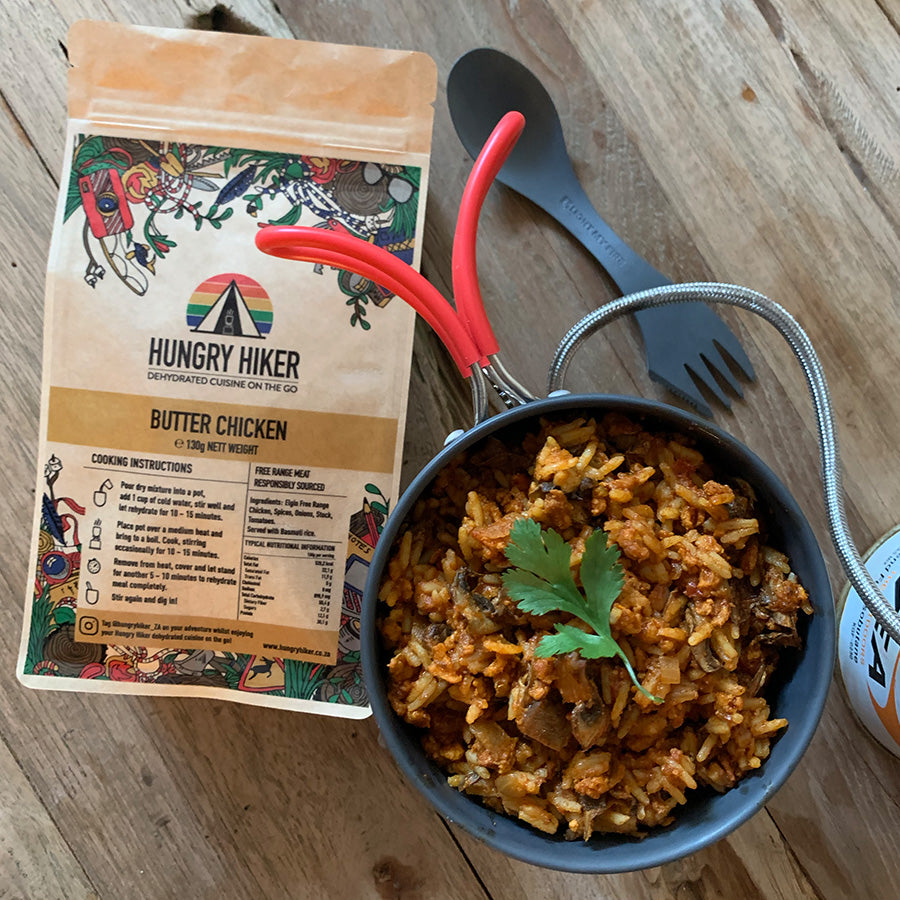 A package of Hungry Hiker Butter Chicken Curry dehydrated food next to a pot of cooked butter chicken curry with Basmati rice, surrounded by a wooden surface and utensils.