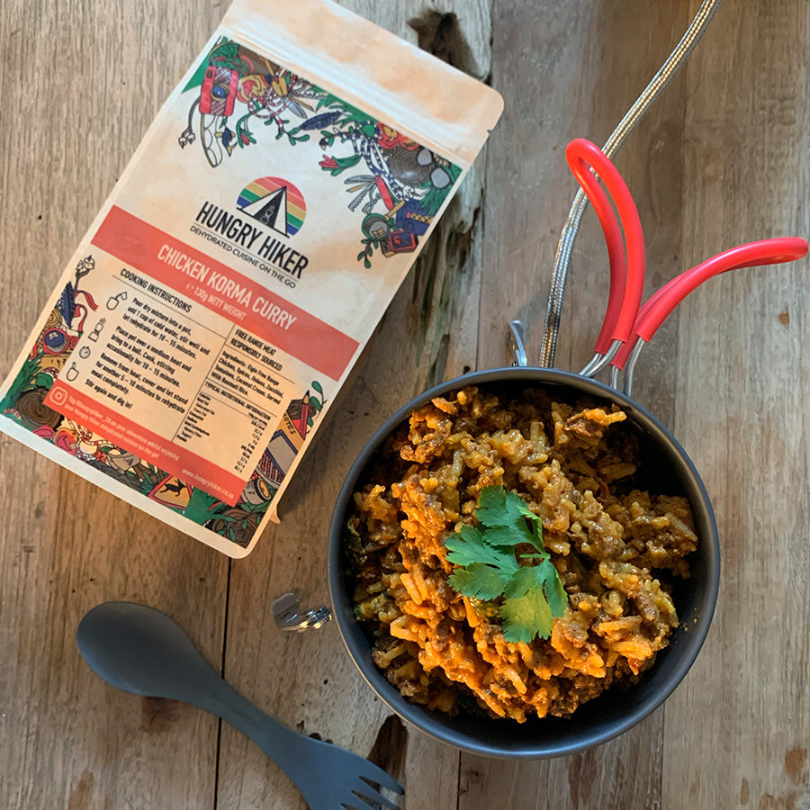 A package of Chicken Korma Curry dehydrated food next to a pot of cooked rice with chopped herbs on top, on a wooden surface with a red spoon and a fork.